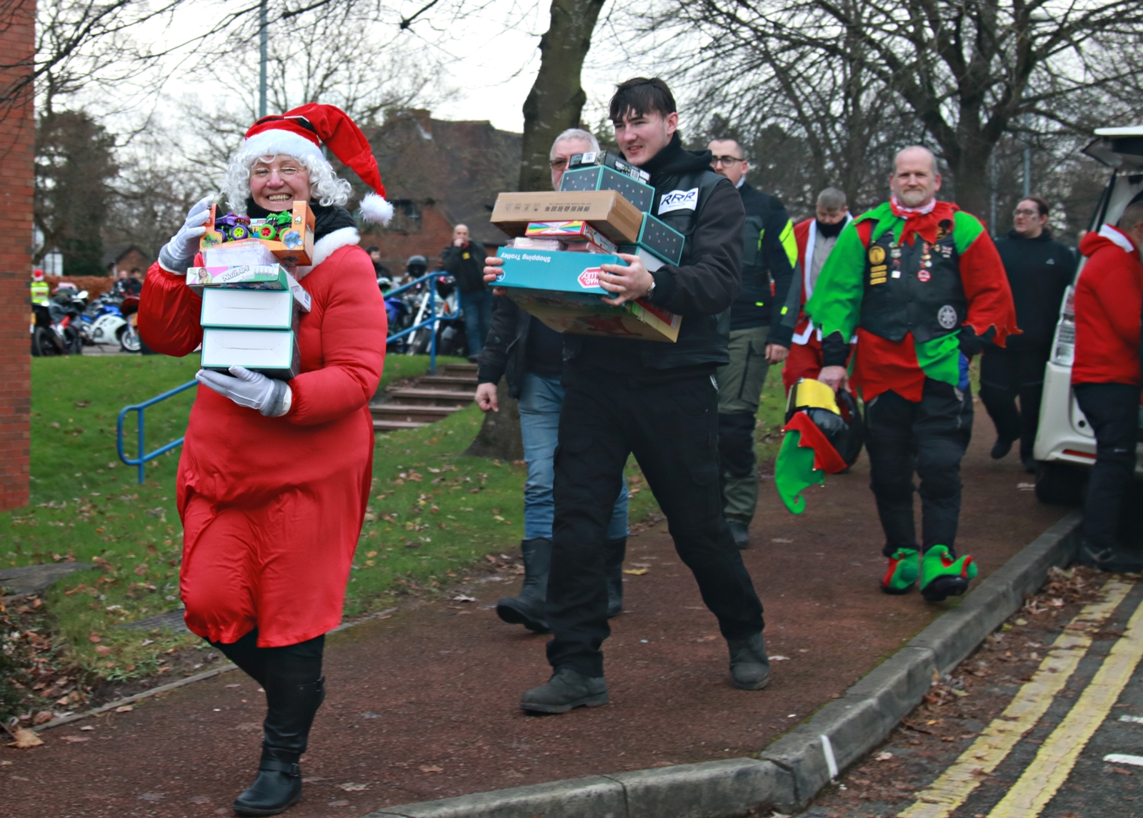 Two people, one dressed as Santa Claus, both carrying a stack of gift boxes