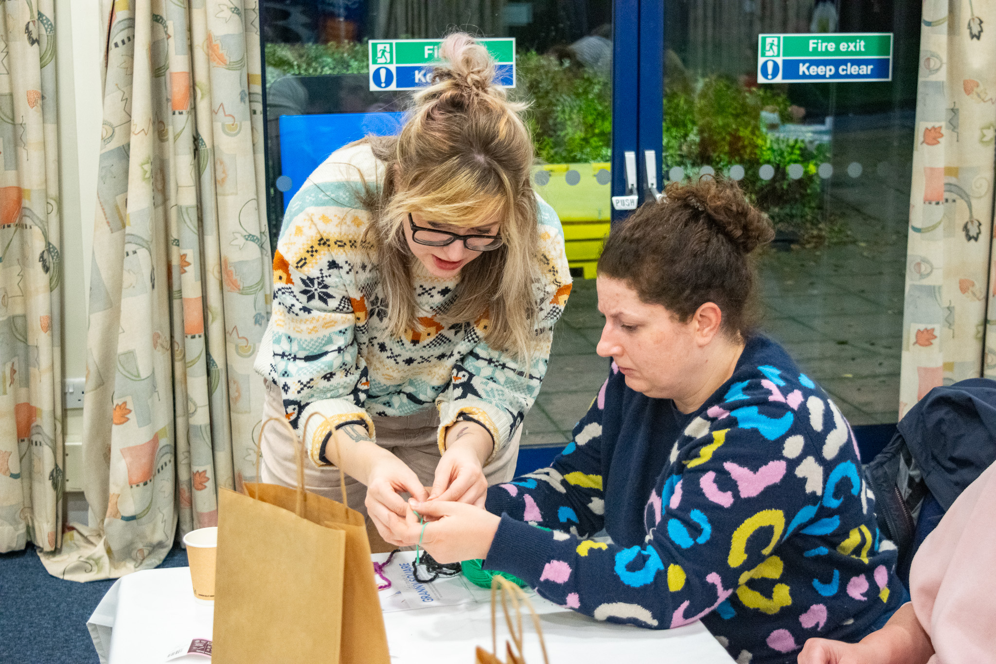 a woman bends down to help another woman with her crochet. 