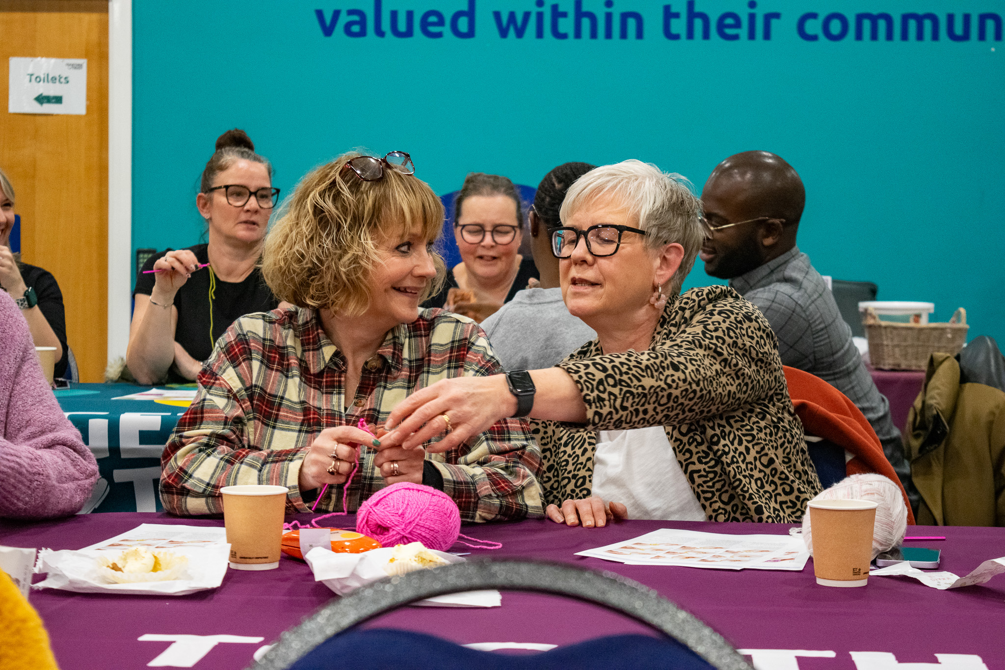 two women smiling at each other as one of them is reaching to help the other with her crochet