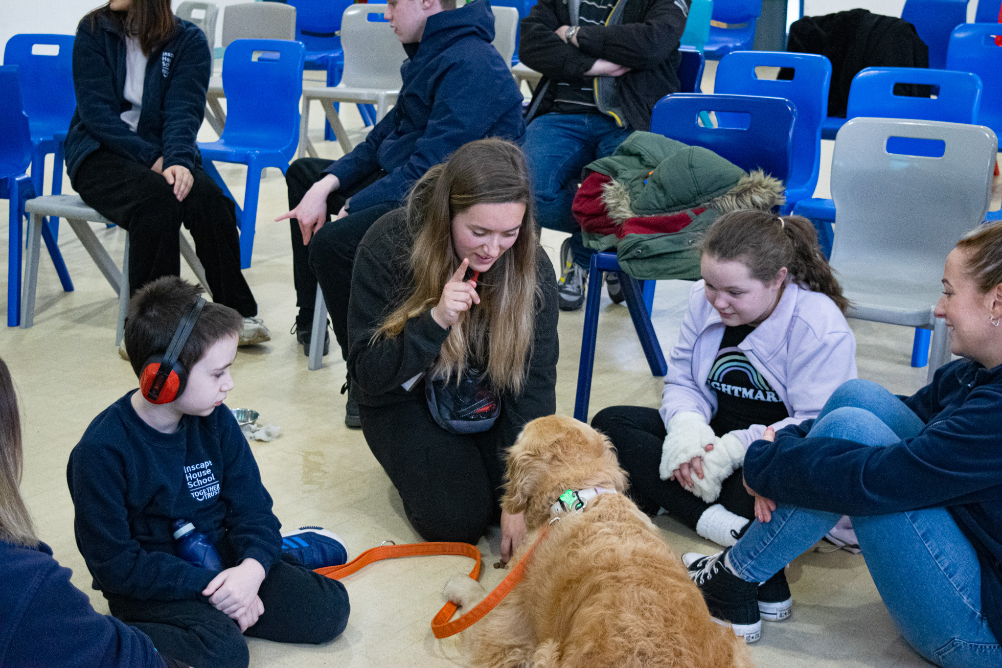 Two young students - one boy with large red headphones and a girl - watch as Laura interacts with Summer, a golden retriever.