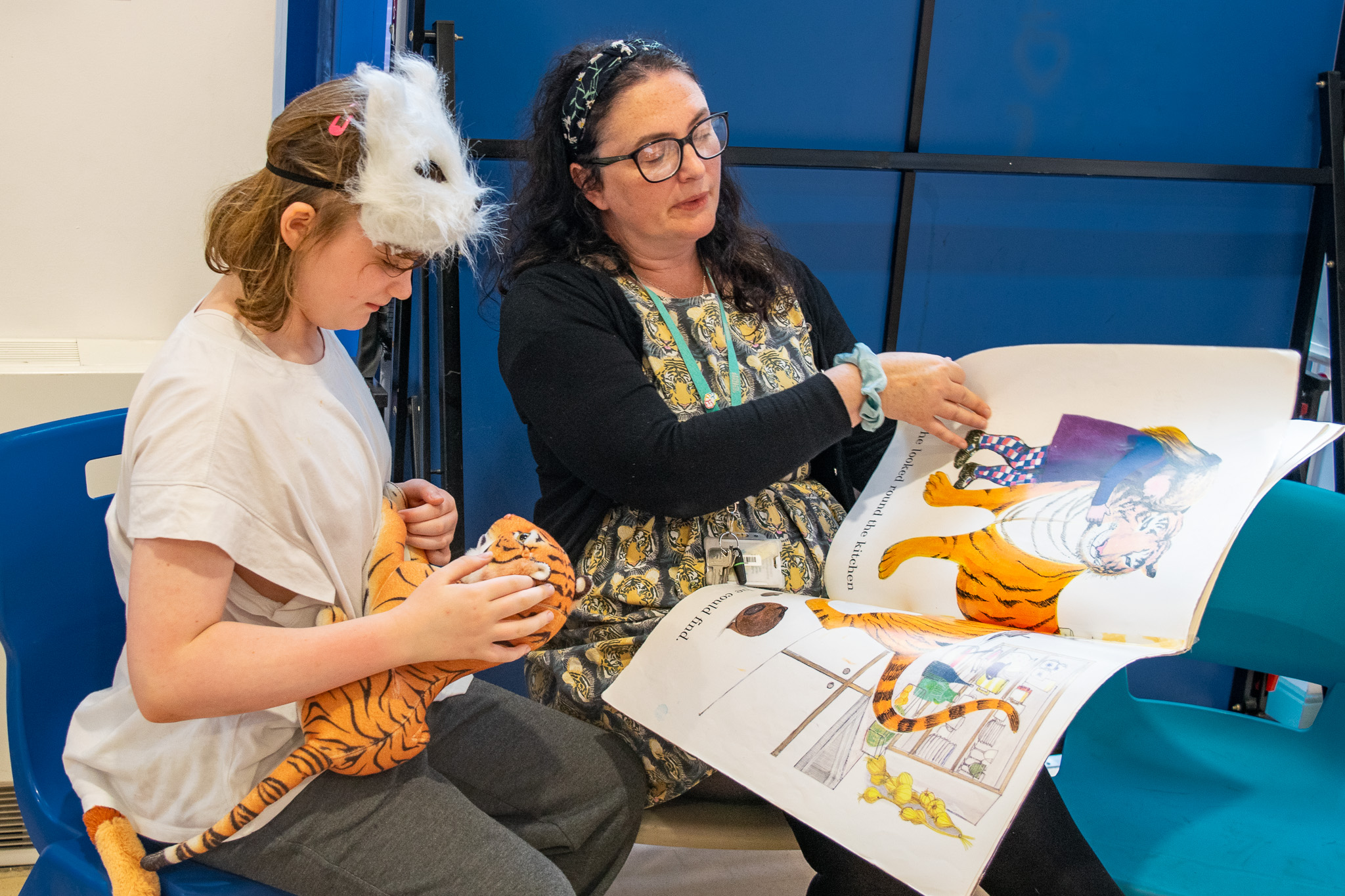 a young girl wearing a white fox mask and holding a tiger toy is sat next to a teacher reading a book about a tiger