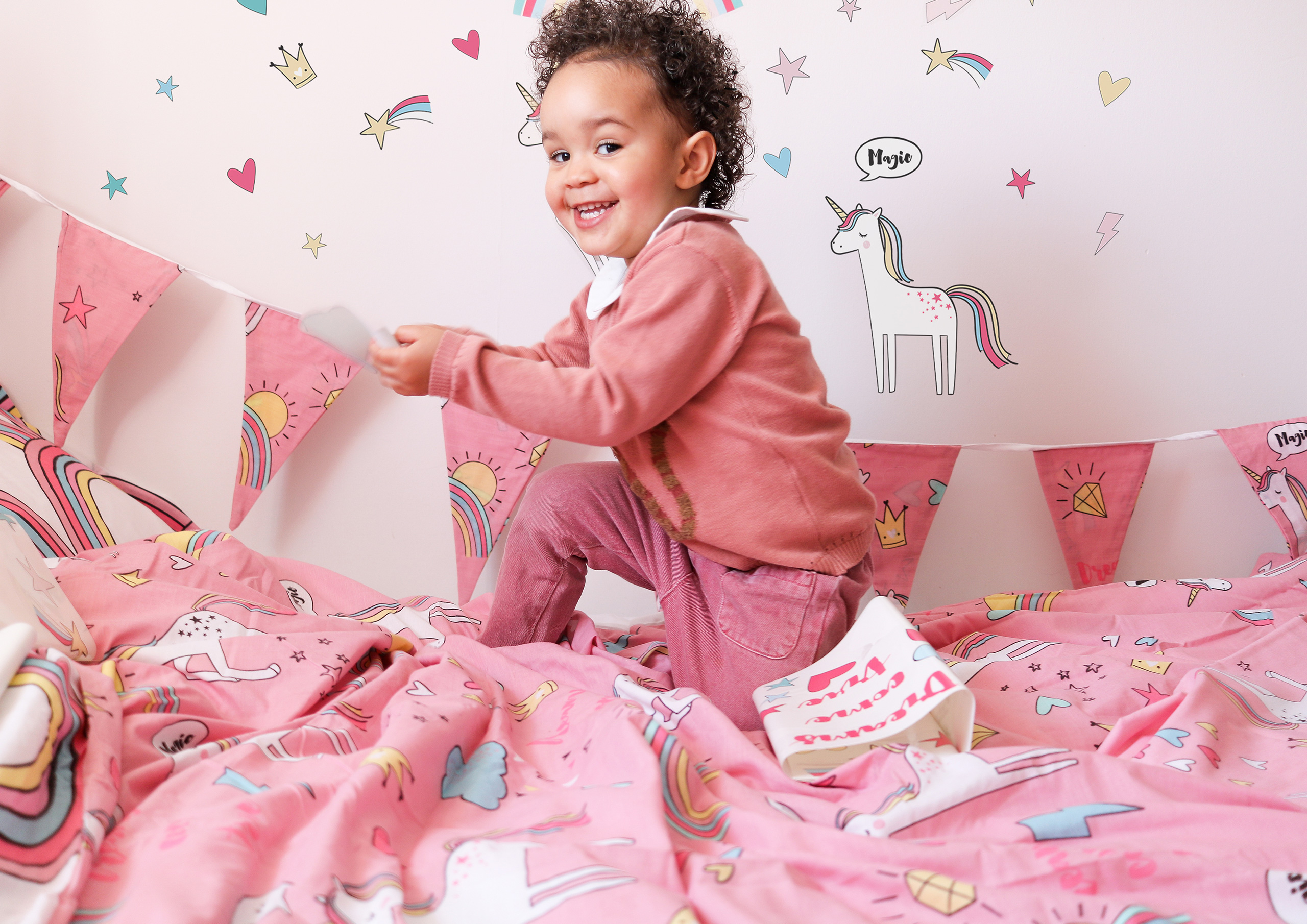 Young girl jumping on her bed. She has matching bunting, wall stickers and bedding in pink with unicorns on. 