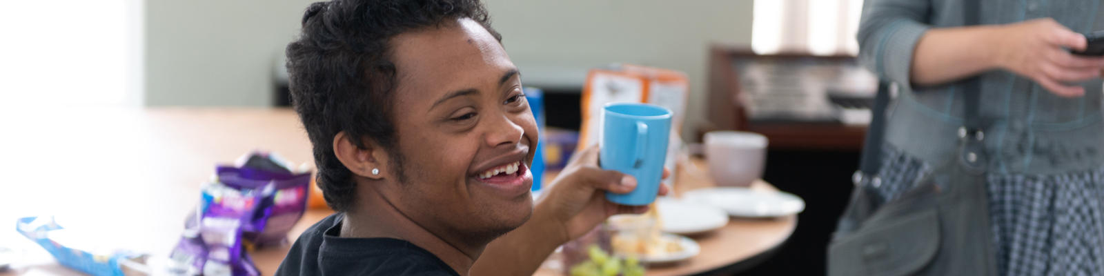 Young man with down syndrome smiling as he is holding a cup. He is sat down at a table. 