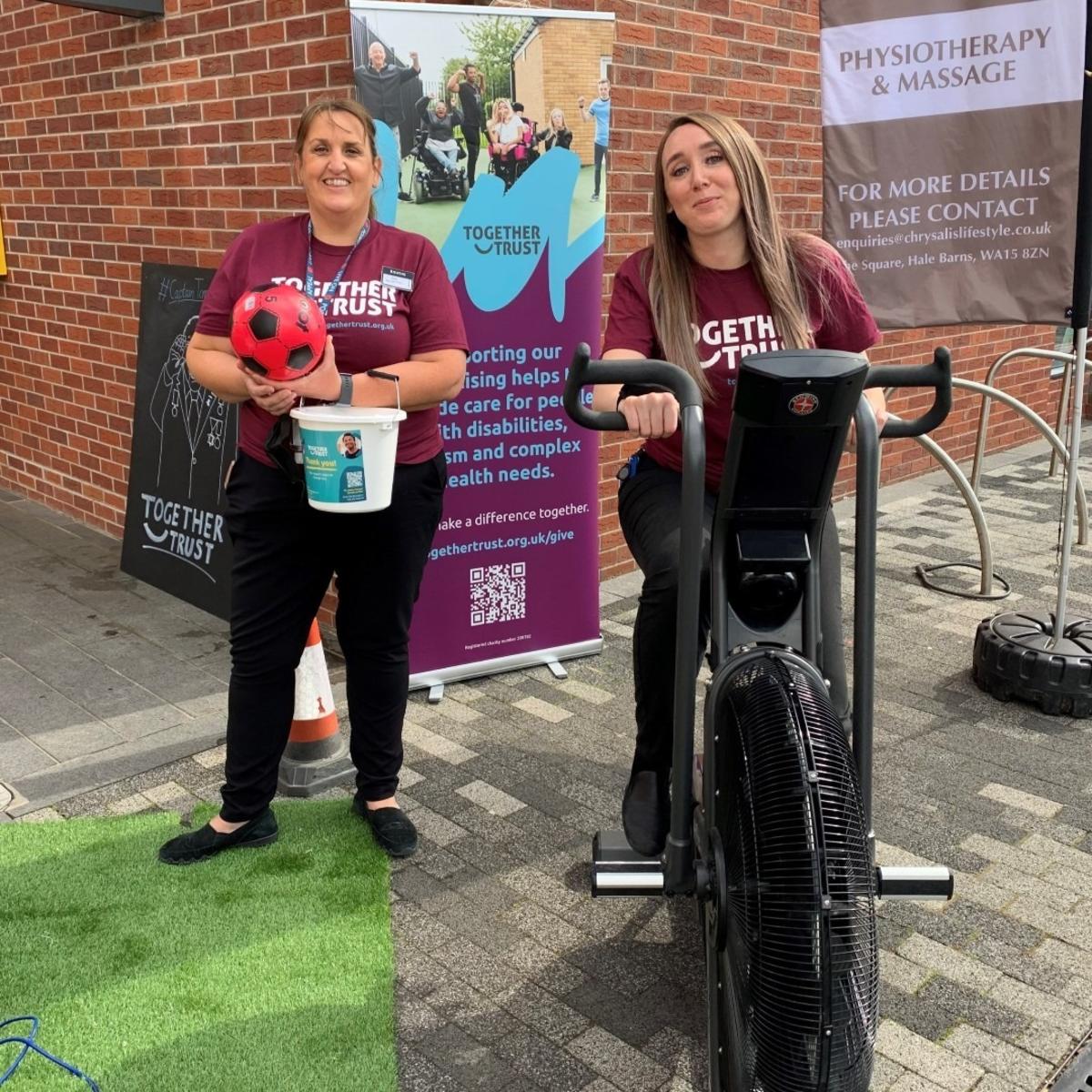 A team member from Booths holding a collection bucket next to another sat on an exercise bike