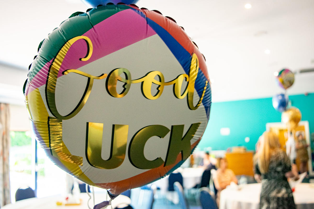 Balloon that reads "Good Luck" floating in a busy room where people are gathering to watch a ceremony. 