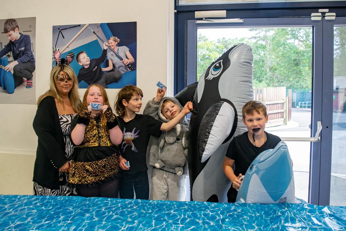 A teacher wearing a zebra costume stands next to a group of 4 students: a girl dressed like a leopard, a boy wearing an axolotl tshirt, a boy wearing a bunny hoodie and a boy holding a dolphin. 