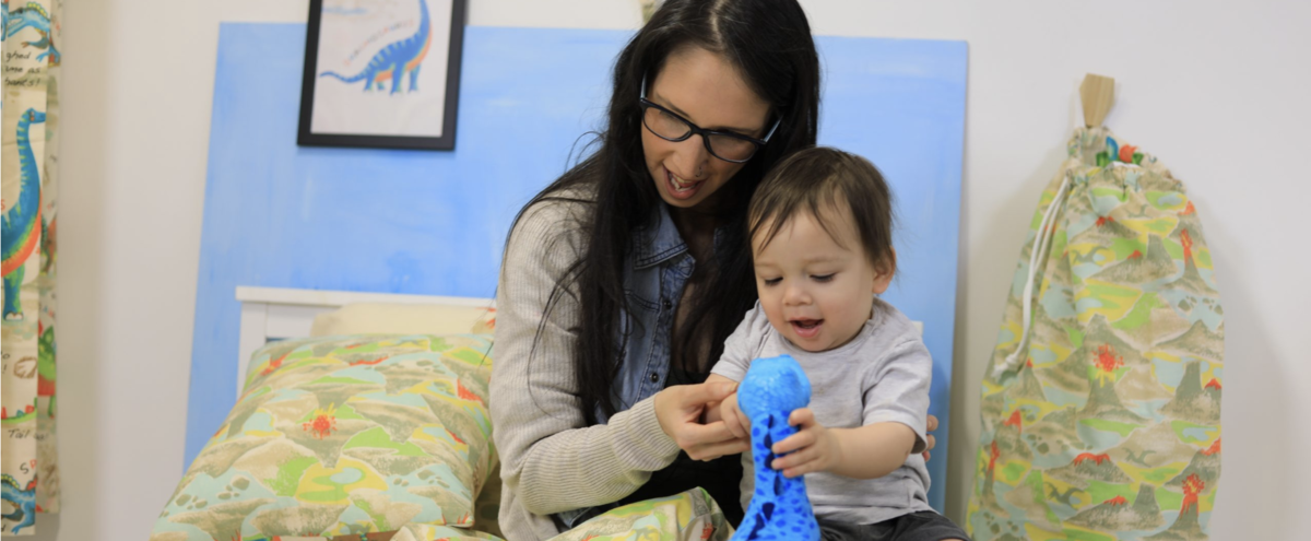 A woman holding her baby as they play with a plush dinosaur. they are sad on a bed with dinosaur bedding. 