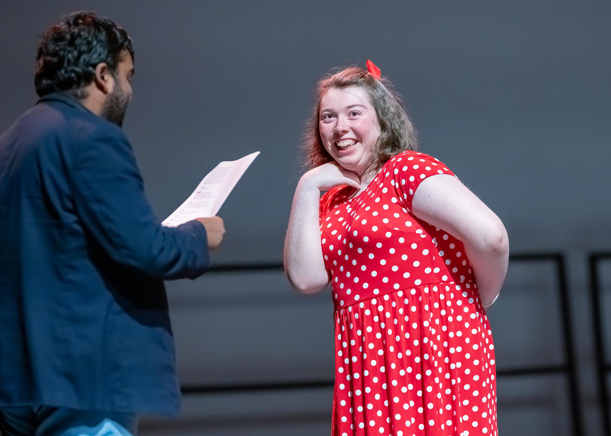 two actors on a stage: A woman in a red dress looking smiling at a man wearing a suit. the man is holding a piece of paper 