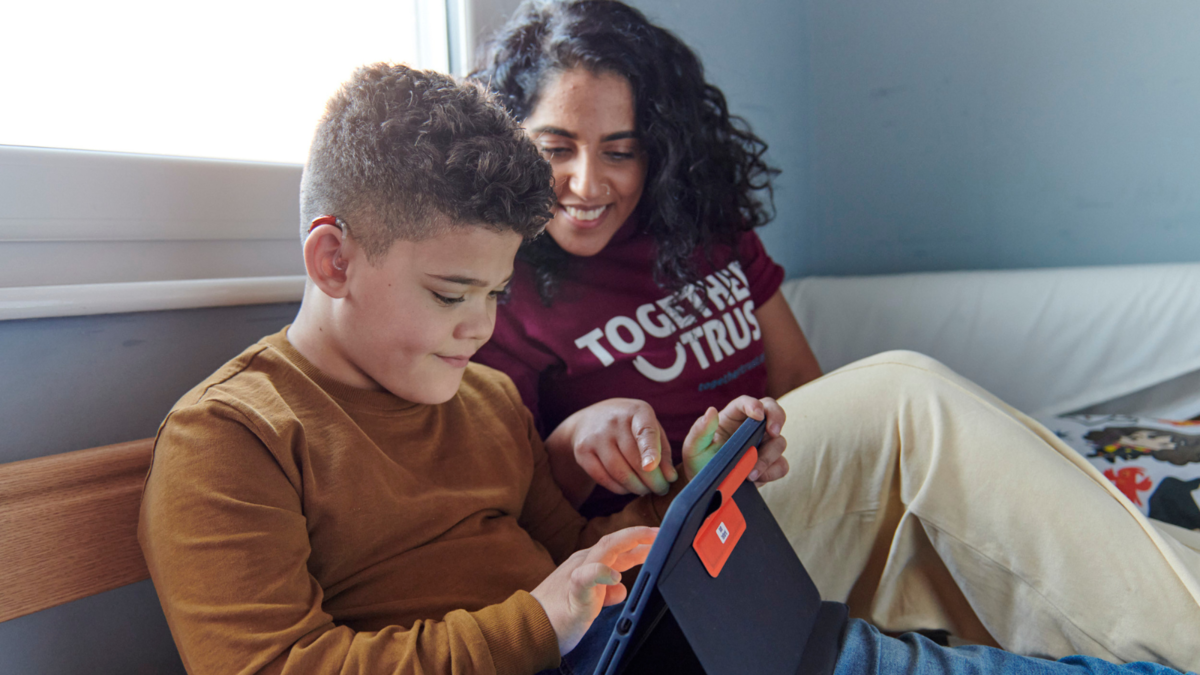 Young boy playing on a tablet in bed. Next to him, a woman wearing a Together Trust t-shirt is helping him choose something on the tablet. 