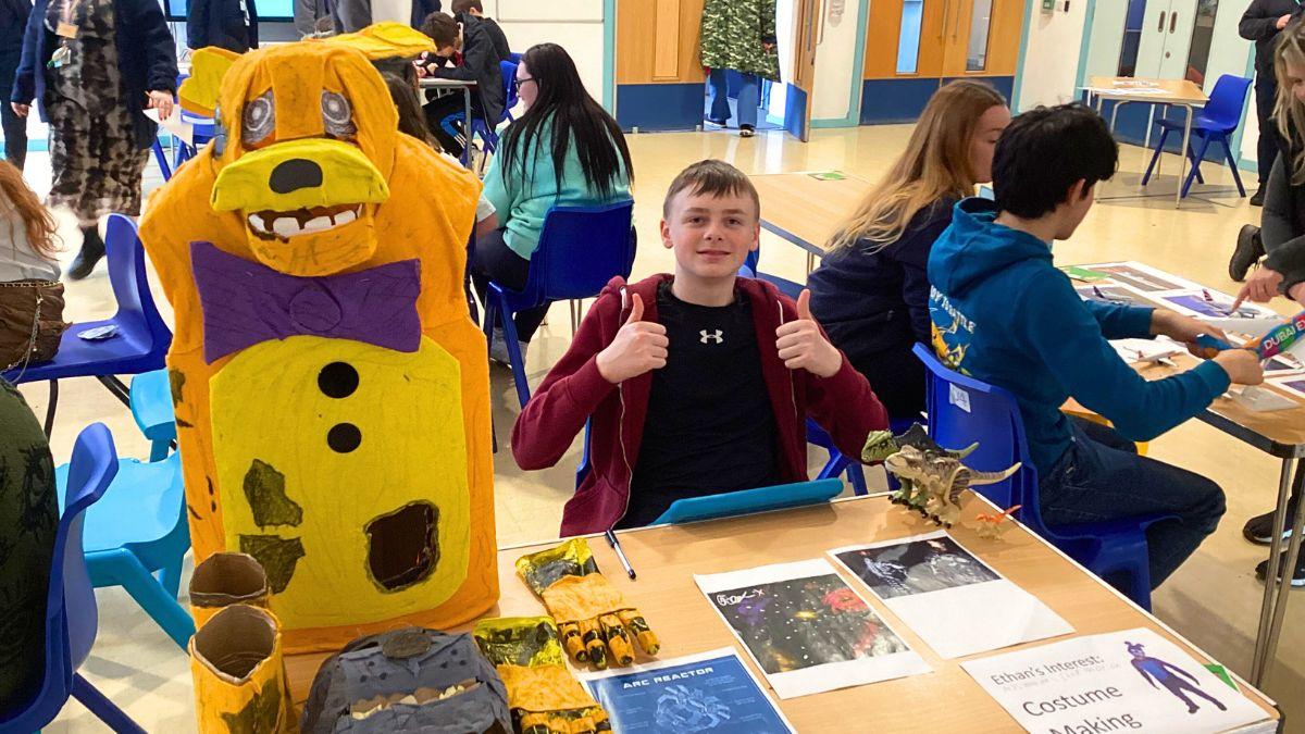 A student proudly running a stall labeled 'Costume Making'. With a genuine smile and a thumbs-up to the camera, he stands next to a vibrant yellow dog costume.