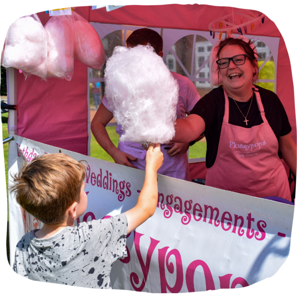 Woman wearing pink apron handing a young boy a big pink candy floss. 