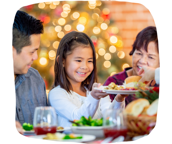 Young girl holding a plate of food sitting down. A man and a woman are looking at her smiling. The room is dressed with christmas decorations.  