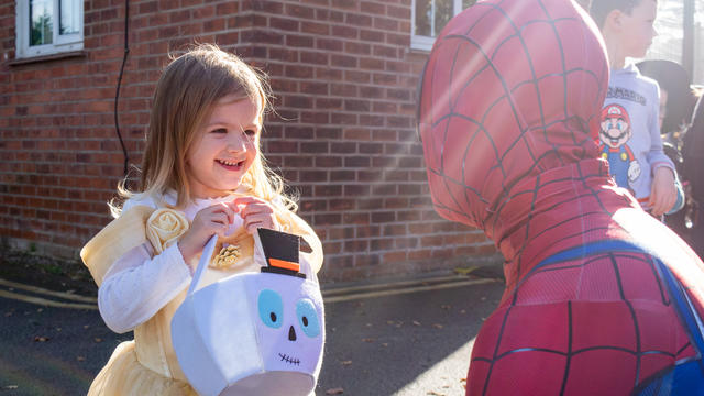 Young girl dressed as a Princess carrying a trick-or-treat bucket, talking to Spider Man.