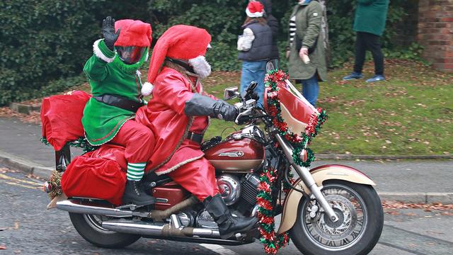 Two bikers on a motorcycle, dressed up as Santa Claus and an elf, waving cheerfully at the camera