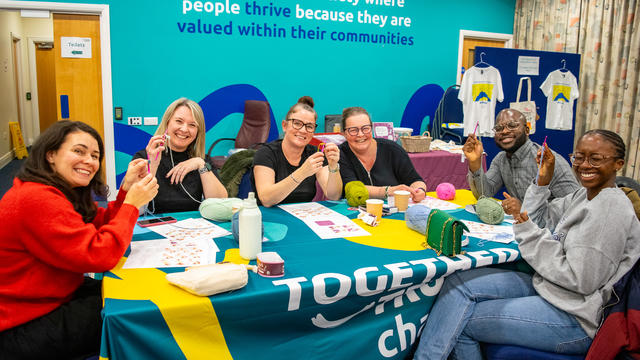 six people sitting around a table smiling into the camera showing off their crochet creations