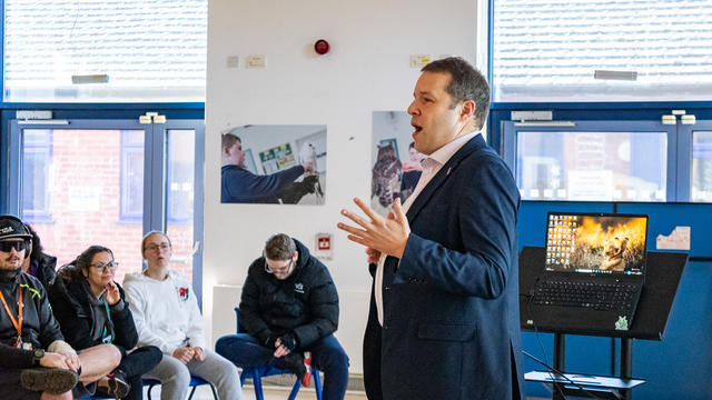 A man in a suit, Mr Morrison, talking to students sat down on chairs in the main hall. 