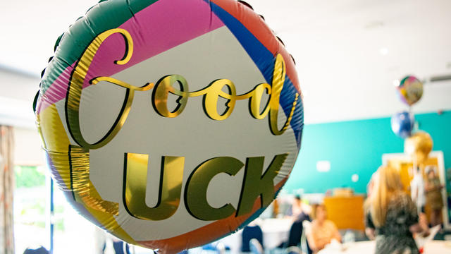 Balloon that reads "Good Luck" floating in a busy room where people are gathering to watch a ceremony. 