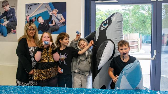 A teacher wearing a zebra costume stands next to a group of 4 students: a girl dressed like a leopard, a boy wearing an axolotl tshirt, a boy wearing a bunny hoodie and a boy holding a dolphin. 