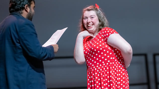 two actors on a stage: A woman in a red dress looking smiling at a man wearing a suit. the man is holding a piece of paper 