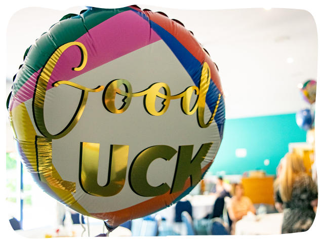 Balloon that reads "Good Luck" floating in a busy room where people are gathering to watch a ceremony. 