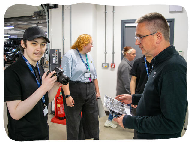 Teen boy holding a professional camera smiling. He is being shown the controls by the photographer. 